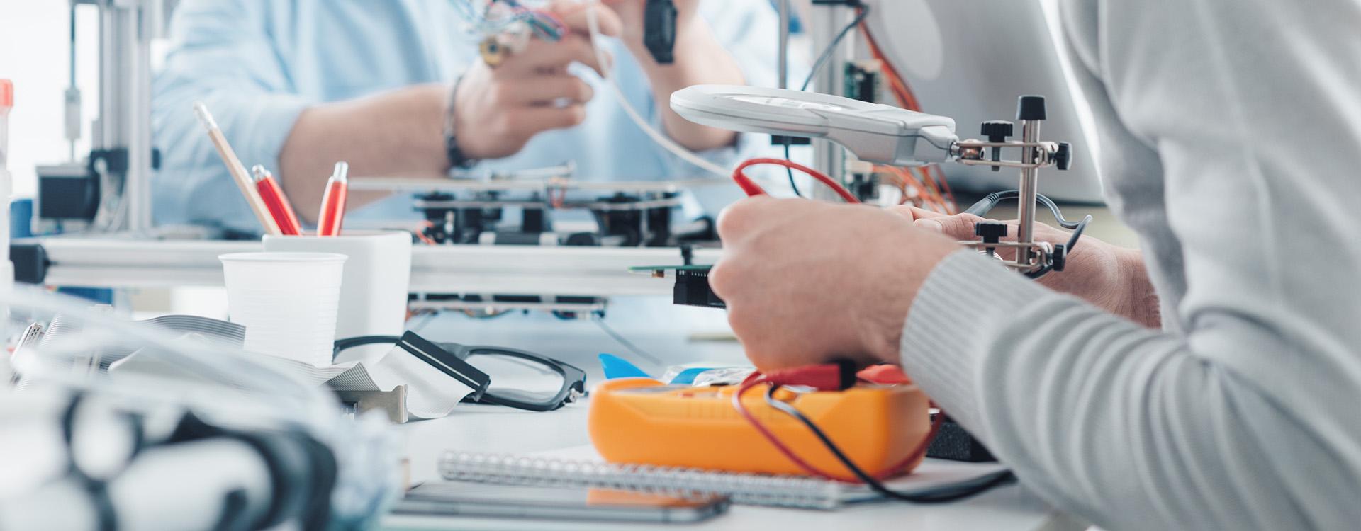 Engineering students working in a lab, a student is using a voltage and current tester, another student in the background is using a 3D printer